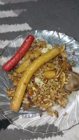 The video shows a close-up view of a plate of food. The plate is clear and has a decorative edge. On the plate, there is a pile of fried rice with what appears to be egg and some other ingredients. Two sausages are placed on top of the rice: one is red and the other is yellow. A fork is visible, actively mixing the food on the plate. To the left of the plate of rice, another plate is visible, this one amber-colored and holding what looks like pink crackers or puffed snacks. The background is blurry but appears to be a carpeted surface with a striped pattern. The lighting is somewhat dim, suggesting an indoor setting. The camera is steady and focused on the food.