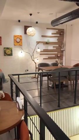 The video shows an interior view of a cafe, taken from an elevated position on a mezzanine level. In the foreground, a black metal railing is visible, separating the mezzanine from the lower level.

On the mezzanine level, there is a dining area with a wooden table surrounded by chairs. The table is set with what appears to be a small vase of dried flowers and a dark circular object, possibly a coaster or small plate. The chairs are upholstered in a dark grey fabric. Behind the dining area, the wall is decorated with wooden planks arranged in a horizontal pattern and a textured beige wallpaper. There are decorative light fixtures and artwork on the walls, including a circular piece with a yellow and orange design and a branching decorative element with small lights.

Looking down to the lower level, there are more seating areas. A large potted plant is prominent, situated near a set of wooden chairs and tables. The chairs have a distinctive patterned upholstery with pink and orange accents. A tall, narrow lamp with a yellow glow stands near the stairs on the lower level.

Through a large window or glass partition on the lower level, reflections and views of the outside or another part of the cafe are visible. People can be seen through the glass. One person, wearing a light-colored garment, appears to be walking or standing. There are also shelves with decorative items, possibly cakes or pastries, visible on the right side of the lower level. The overall ambiance suggests a modern and stylish cafe.