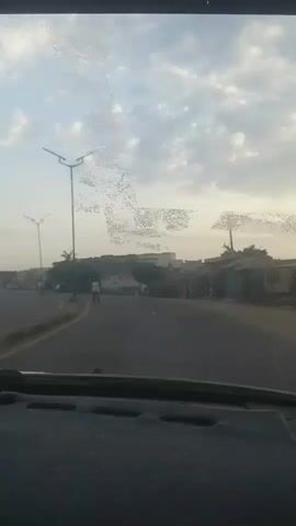 This video shows a view from inside a car looking out through the windshield. The car is driving on a paved road. On the left side of the road, there is a sidewalk with a lamppost and some people walking. On the right side of the road, there are buildings and some vehicles parked. The sky is cloudy. The scene appears to be a street in a town or city.