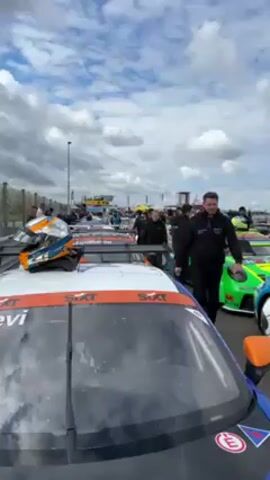 The video shows a racetrack on a partly cloudy day, featuring a row of parked race cars. The camera is positioned close to a white, blue, and orange race car with “Sixt” branding and the name “Levi” on its windshield. A helmet, matching the car’s colors, rests on the roof. Several individuals in team attire are walking among the cars. In the background, there are more cars, tents, and spectators, all suggesting a racing event.
