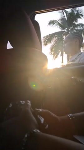 The scene is inside a car, with the sun shining brightly through the window.  A person is visible outside the car, seen from the window, with palm trees in the background. The person has their hair in braids, is wearing a light-colored shirt and appears to be looking towards the camera. Inside the car, two hands are visible. One hand is holding something dark and unclear, while the other is resting on top of it. The hands are adorned with beaded bracelets. The silhouette of a person sitting in the passenger seat is also visible.