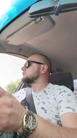 A man with a shaved head, beard, and sunglasses is driving a car. He is wearing a white t-shirt with a pattern of motorcycles and a black crossbody bag. The sun is shining, and there are trees visible outside the car window. The car's interior is visible, including the dashboard and rearview mirror. The man looks towards the right side of the car, then ahead. He adjusts something on his wrist, revealing a watch. He then turns his head slightly to the left and looks forward again.
