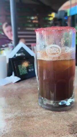 The camera pans slowly around a glass of beer on a table in an outdoor restaurant. The beer is dark brown and bubbly, and the rim of the glass is coated in a bright red powder.  A logo on the glass, blurred by condensation, is readable as "Carta Blanca."  Other objects in the scene include a potted plant and a small sign behind the drink, and a partial view of one or two other patrons in the background. The table has a speckled beige surface.  A second, similar drink is visible out of focus at the left edge of the frame.