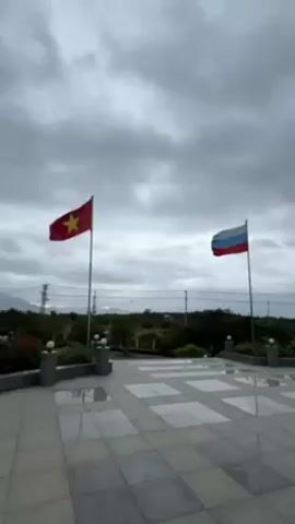 The video shows a gray, cloudy sky above a paved area with the Vietnamese and Russian flags waving side-by-side.  The flags are on poles in front of a view of low vegetation. The camera then pans to show a large stone monument of sailors aboard a ship, viewed from below.