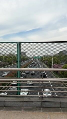 The video shows a view from a bridge overlooking a highway. There is traffic on the highway, including cars and trucks. A green mesh fence separates the viewer from the highway. The sky is cloudy with some blue patches. Buildings and trees are visible in the background.