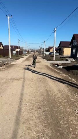 A young boy on a green and black mountain bike rides towards the camera on a dirt road in a rural or suburban area. He is wearing a camouflage jacket and pants, a cap, and gloves. The sky is clear and blue. Utility poles with wires line the road, and houses with brown roofs are visible on either side. The road is dry and dusty with patches of gravel. The boy smiles and says "Mama" as he gets closer to the camera.