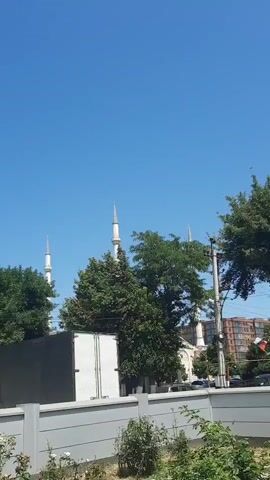 This video appears to be a recording from a moving vehicle looking towards a mosque with multiple minarets. The sky is clear and blue, indicating a sunny day. Lush green trees partially obscure the mosque's structure. In the foreground, a grey fence and some vegetation are visible. A white truck or van is parked near the mosque. In the background, a building with a reddish-brown facade can be seen. The audio consists of an Islamic prayer being recited.
