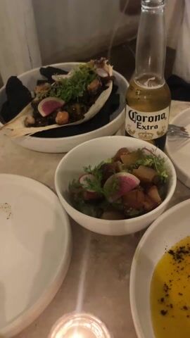 A handheld video shows a restaurant table with multiple dishes, including salads with vegetables and possibly seafood, and a yellow sauce with black specks. A bottle of Corona Extra beer sits near one of the dishes. The camera moves around the table, revealing a mostly empty plate and then pans to other diners, showing them seated and a waiter walking by with a basket. The video ends with a woman with dark hair looking down at her phone. The restaurant appears to be an open-air venue with a wooden-slatted roof. Loud music plays throughout the video.