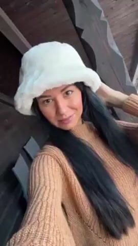 A woman wearing a white furry hat and a tan-colored sweater poses for a selfie. She is standing on what appears to be a porch or balcony with wooden beams overhead. Her hair is long, straight, and black. She has her right hand touching the side of her hat as she looks at the camera and smiles slightly. The background shows glimpses of trees and potentially an outdoor environment. The lighting is soft and natural, contributing to a calm atmosphere.