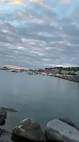 The video opens with a view of a harbor at dusk, showing several boats floating on calm water. The sky is filled with textured clouds, tinged with the orange glow of the setting sun. In the foreground, large rocks line the shore. The camera pans right, revealing a stone pier leading into the harbor. A white statue stands at the end of a rocky outcrop adjacent to the pier. As the camera moves further along the pier, a streetlamp with security cameras becomes visible. The camera then rotates to show a woman standing on the pier, facing the camera. She is smiling. She is wearing a beige top and a denim jacket. The background reveals the harbor and the buildings along the waterfront.