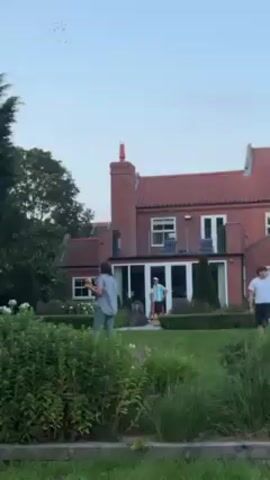 Two people are standing in a garden in front of a brick house. The person in the foreground is gesturing with their right arm. The other person is walking in the background. They are both wearing jeans. The brick house has a red roof and white windows. There is a smaller wooden building next to the house. The garden is full of flowers and greenery. The sky is overcast. The person in the foreground says, "Right, four four four four for one."