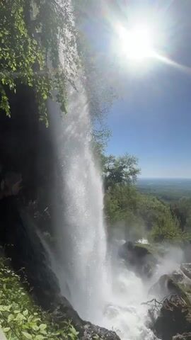 The video shows a tall waterfall under a bright blue sky with a visible sun flare. The waterfall cascades down a rocky cliff covered in greenery. At the bottom of the falls, the water churns into a misty spray. Beyond the waterfall, the view opens to a vast, green landscape dotted with trees and hills. The overall impression is of a scenic natural environment on a sunny day.
