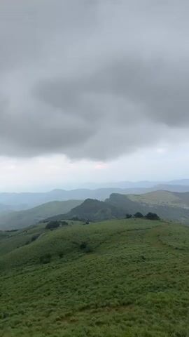 The video shows a wide shot of a mountain range on an overcast day. The foreground is a green, grassy hillside, while the background consists of a series of rolling hills and mountains, with a body of water visible in the distance. The sky is filled with heavy, grey clouds, but there are patches of blue sky breaking through. The camera slowly pans from left to right, revealing more of the landscape. The overall impression is one of natural beauty and tranquility, despite the cloudy weather.