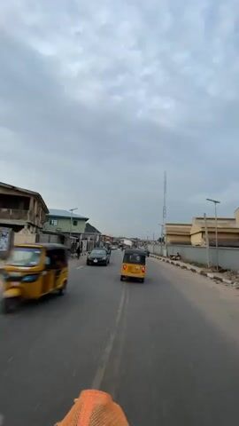 The video shows a view of a road with traffic on a cloudy day. The perspective is from behind a person on a motorcycle or similar vehicle moving down the road. Several vehicles are visible: a black car, a yellow "Keke Napep" (a type of auto-rickshaw common in Nigeria), and several motorcycles. There are buildings of various heights and architectural styles on either side of the road. A tall communication tower is visible in the distance on the right side of the road. People are walking along the sidewalks. The road is paved with yellow lines dividing the lanes. The general environment suggests an urban or suburban setting in a developing country.