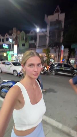 A fair-skinned woman with blonde hair in a low bun, wearing a white tank top and light blue bottoms walks along a street at night, holding the camera. She passes a red vehicle decorated with lights. Street lights and store signs illuminate the scene. Other people are visible on the street, and parked cars and motorbikes line the curb.