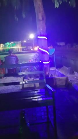 The video shows an outdoor seating area at night. There are people sitting at tables, and a tree is decorated with blue LED lights. The scene is lit with various colored lights, including blue, green, and white. There is a bench in the foreground, and a bottle is sitting on the ground near it. The camera slowly pans to the left.