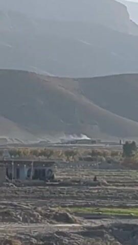 The video shows a landscape with mountains in the background and a flat, rocky terrain in the foreground. The ground appears to be mostly barren, with some patches of vegetation visible. The sky is clear and blue. The camera is moving, likely from a vehicle on a road.
