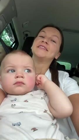 A woman holds a baby in a car. The woman is singing with her eyes closed. The baby is looking up and to the left with a neutral expression. Another person is visible in the background in the car. The scene is lit and the car appears to be in motion, suggested by the motion blur in the background.
