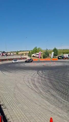 The video shows a drift competition on a sunny day. Two cars, a yellow sedan and a white sedan, are drifting around a course marked by orange cones. A crowd of spectators is visible in the background, as well as tents and banners. The ground is paved with interlocking stones, and there are tire marks on the surface from the drifting. There are also a few other cars parked in the background. A fire extinguisher is visible in the lower left corner of the frame.