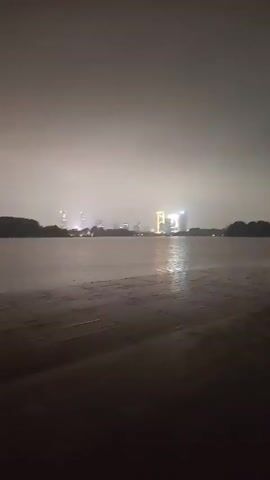 A video shows a body of water with a cityscape in the background. The sky is dark and hazy, with lights from the city visible. The water appears calm, with a slight ripple. In the foreground, a sandy or muddy shore is visible. Two figures, appearing to be people, stand near the water's edge. The overall atmosphere is dimly lit, suggesting nighttime or heavy fog.