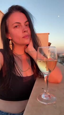 A woman is taking a selfie on a balcony at sunset. She has long, dark hair and is wearing a black tank top and jeans. She is holding her head in her hand and looking at the camera with a thoughtful expression. To her right, on a ledge, is a glass of white wine. The sun is setting, casting a warm, golden glow on the scene. The sky is a clear blue with a hint of pink and orange from the sunset. A small moon is visible in the upper right corner of the frame. In the background, there are buildings and a hint of the landscape of Katzrin, Israel. The overall mood of the image is peaceful and serene.