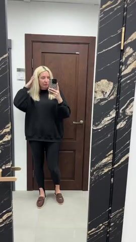 A blonde woman stands in front of a mirror, taking a selfie. She is wearing a black sweatshirt and black leggings. The woman has blonde hair and is wearing leopard print nail polish. She is holding a phone with a leopard print case. In the background, there is a wooden door with a gold handle and a large mirror with a black and gold marble frame. The floor is tiled.