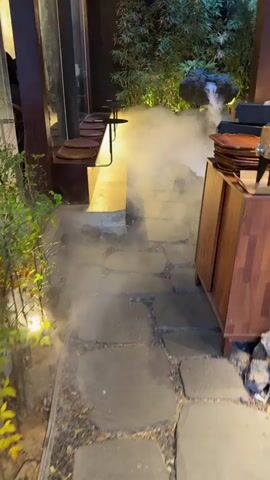 The camera pans up to reveal a tranquil outdoor setting at a restaurant or bar, the atmosphere enhanced by the presence of artificial fog drifting low to the ground. The space features a stone pathway leading to a small rock formation, emitting a trickle of water and surrounded by lush green foliage. Ambient lighting illuminates the scene, casting a soft glow on the plants and the fog. A wooden cabinet stands to the right of the path, topped with a stack of decorative plates. To the left, there's an elevated seating area with a row of chairs along a counter. The setting gives off an inviting, peaceful ambiance, likely designed to create a unique dining or social experience.