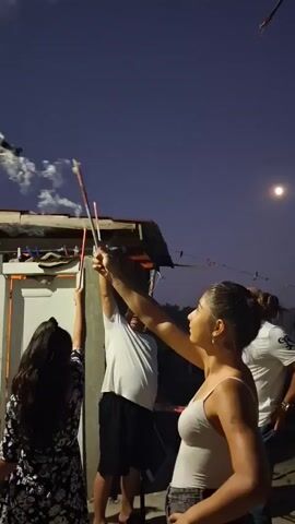 Three people are standing outdoors at night, holding up sparklers. A fourth person is visible in the background. The sky is dark blue with a full moon. There is a building with a corrugated roof behind the people. The sparklers are emitting sparks and smoke. The person in the foreground, a woman with her hair tied up, is looking up at the sparkler she is holding. Another woman, with long dark hair, is to her left, also holding a sparkler. A man in a white t-shirt is in the middle, also holding a sparkler, and a fourth man in a white t-shirt is to the right. The overall atmosphere appears festive.