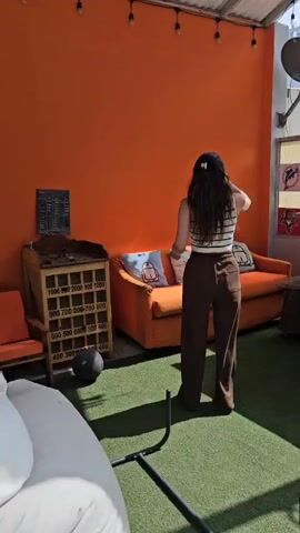 The video shows a small outdoor gym area on a rooftop. It opens on a woman with long hair standing on an artificial grass surface. Behind her is an orange wall, a small orange couch with two throw pillows, and a wooden cabinet-like structure with slots for holding objects. The video then pans to the right showing a wooden bar, two painted doors, one blue and one orange, propped up behind it. The camera continues to pan, showing a concrete floor with a blue punching bag hanging from the wall and a blue door to what appears to be a small storage room. The final pan reveals a black metal fence with a weight lifting bench and bar behind it. The scene is bright with natural light.