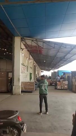 The video shows the exterior loading bay of a warehouse. Several men are visible, one is wearing a green jacket and standing in the middle of the frame. A man in a black shirt walks towards the loading bay doors. A green cargo truck with “BESAR, RT CARGO” on its side drives up to the doors. A man on a blue scooter drives away from the warehouse area. The sky is visible above the warehouse overhang. There are several trees outside the warehouse building.