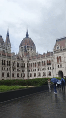 Pedestrians with umbrellas walk past Hungarian Parliament on overcast day
