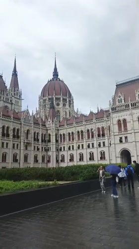 Pedestrians with umbrellas walk past Hungarian Parliament on overcast day