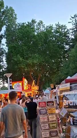 The video captures a lively scene at an outdoor event, possibly a fair or festival, during dusk. A crowd of people is visible, walking and interacting amidst various stalls. Dominating the center and right side of the frame is a stall with a white canopy decorated with strings of lights, advertising "Deutsche Küche Landmetzgerei mit 9 Pfannen." Several patrons are gathered around this food stall, with some appearing to be ordering or purchasing food. To the left of the food stall, another stall with red and white checkered tablecloths is partially visible, also appearing to be a food vendor. In the background, a larger structure with bright lights and signage, possibly an amusement ride or another attraction, is illuminated. Tall green trees form a canopy overhead, suggesting the event is set in a park-like environment. Several people are in the foreground, walking away from the camera, showcasing casual summer attire. The overall atmosphere is one of a bustling public gathering with food, entertainment, and a festive ambiance.