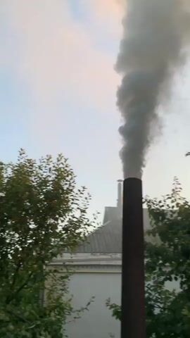 The video shows a close-up of a chimney with dark smoke billowing out of it against a pale sky with some clouds. The chimney is made of dark, possibly rusted metal, and appears to be cylindrical. Green trees are visible on the right side of the frame, and the roof of a building is visible on the left side, behind the chimney. The smoke is thick and dense, rising high into the air and dissipating. The overall impression is one of industrial activity or a fire.