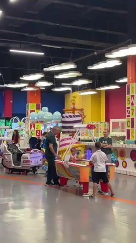 The video pans across an indoor children's entertainment facility. In the foreground, a foosball table with a soccer ball design is visible. Further back, a soft play area with yellow slides and climbing structures can be seen, along with various arcade machines and kiddie rides, including a train and what appears to be a fire truck ride. Several people are present, including adults and children, interacting with the games and play structures. The environment is brightly lit and decorated with vibrant colors.