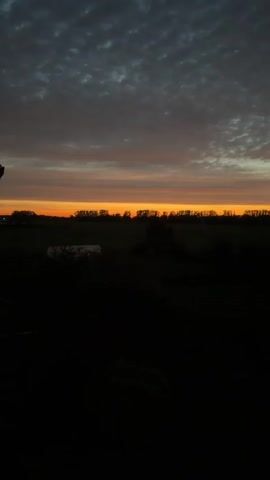 The video shows a sunset over a rural landscape. The sky is filled with dramatic clouds, ranging from dark gray to bright orange and yellow hues. A thin line of orange light marks the horizon, separating the sky from the dark silhouettes of trees and a distant field. In the foreground, a portion of a building, possibly a house, is visible on the left side, with a roof and a section of a wall. The rest of the foreground is very dark, obscuring details of the ground. The scene is still and peaceful, with no visible movement or activity.