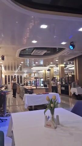 The video shows the interior of a restaurant with a buffet area. Multiple tables with white tablecloths and flower arrangements are visible, and people are seated at some of them. Staff members are walking around, presumably attending to customers. The restaurant is well-lit with a modern lighting system, including colored stage lights. The overall ambiance suggests a formal or upscale dining experience.