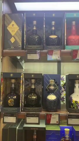 A full shot shows a store shelf with various bottles of Royal Salute whiskey. The top shelf contains six bottles, with the left-most bottle being the Royal Salute 25, followed by two dark blue bottles, a red bottle, and a dark blue bottle on the far right. The bottom shelf also displays six bottles. From left to right, there are a dark blue bottle, a light blue bottle, a white bottle with floral patterns, a pink bottle, and a green bottle. Prices are visible on small white labels beneath some of the bottles. The store appears well-lit.