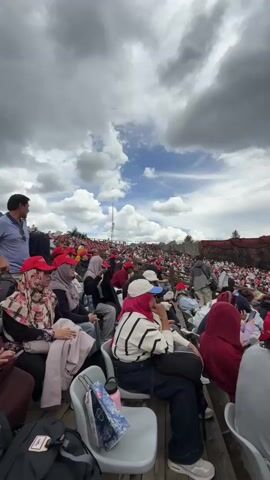 A vertical, medium shot captures an audience seated in an outdoor amphitheater, facing a performance area. The sky above is filled with dramatic, heavy gray clouds interspersed with patches of blue sky and wispy white clouds. The seating appears to be wooden bleachers with individual gray seats. Many audience members are wearing red baseball caps. In the foreground, a person in a white and black striped sweater and a pink hijab is seated, holding a phone. To their left, another person wears a pink and white patterned hijab. Further down the rows, more people are visible, many in red caps, looking towards the performance area. The stage area is a large, reddish-brown structure with a textured surface, resembling rock formations. A few figures can be seen on the stage, and there's a tall, thin pole or antenna in the distance. The overall atmosphere suggests a gathering for an outdoor event or show, possibly in a mountainous or natural setting given the backdrop and the cloud formation.