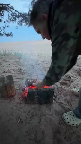A person wearing a camouflage top and white shoes with black dots crouches on the sand next to a small bonfire. The fire is made of logs and is burning with orange flames and white smoke. The person is holding a pine cone and places it into the fire. The sand is light brown and a green plant is visible in the background on the left. Beyond the sandy area, there is a body of water and a light blue sky.