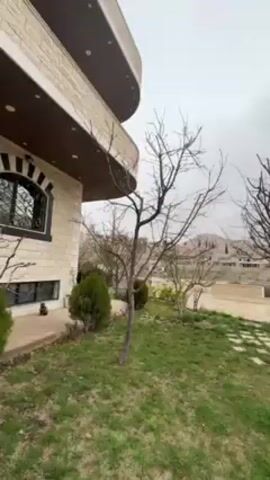 The video presents a view of a lawn on an overcast day. The grass is green, punctuated by square stone pavers arranged in a pathway. Several bare-branched trees stand on the lawn. To the left, part of a two-story stone building with a curved balcony is visible. Across the lawn, a low retaining wall is topped with vegetation and planters. Beyond the yard, a hilly landscape dotted with buildings is visible. The sky is uniformly gray, indicating an overcast weather condition.