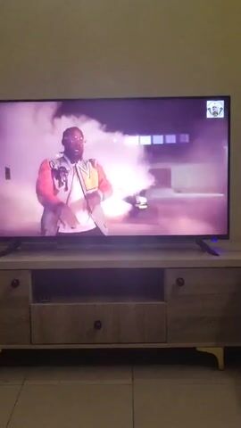 Here's a description of the video:

A television is playing a music video featuring a man with a beard, wearing sunglasses and a black suit. He is surrounded by dancing women in dark-colored swimwear. The scene transitions to the man in a car with another man, both drinking from cups and wearing patterned shirts. The TV is sitting on a light wood-grain TV stand with drawers and open shelving. The floor is tiled.