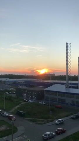 This is a vertical, eye-level shot of a sunset over an industrial area. The sun, a bright orange orb, is positioned in the center of the frame, partially obscured by wispy clouds tinged with orange and pink. The sky transitions from a pale blue at the top to a warm, golden hue near the horizon.

In the foreground, a street curves through the scene, with several cars parked and driving on it. The cars are of various colors, including silver, white, red, and black. A pedestrian crossing is visible on the left side of the street.

Behind the street, there are several industrial buildings. One building in the mid-ground has a dark facade with many windows and appears to be a warehouse or office complex. To the right of this building is a larger structure with a blue and white roof, and a tall, industrial smokestack with a metallic lattice structure rises prominently to the right of the frame. Another, shorter smokestack is visible to the far right. The buildings are set against a backdrop of green trees, and in the distance, faint outlines of urban buildings can be seen under the setting sun.

The overall impression is one of a serene end to the day in a semi-urban industrial setting. The time of day is clearly sunset, indicated by the low angle of the sun and the warm colors in the sky. The weather appears clear, with only a few scattered clouds. There is no visible text in the media.