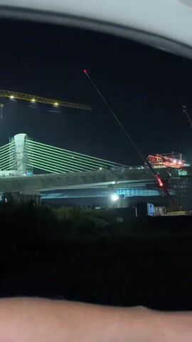 A night-time view from inside a vehicle looking out a window at a bridge construction site. A large yellow crane is visible on the left, and a red construction crane is in the center. The bridge structure, illuminated by green and white lights, spans across the frame. There are scaffolding and building materials around the bridge. The sky is dark. The bottom of the frame shows a person's arm and hand in the foreground, suggesting the viewer is a passenger. The video appears to be handheld and slightly shaky, with a faint, indistinct sound.