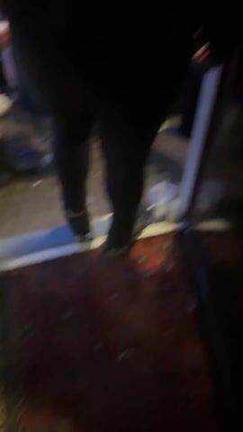 The video starts with a close-up of someone's feet stepping off a rug-covered step into an area with litter. The camera moves upwards, showing a crowded street scene at dusk or dawn. Many people are visible, mostly standing and conversing, with buildings lining the street and strings of colorful lights overhead. A sign for a "Bar" is visible on one of the buildings. The sky is a deep blue, suggesting either early morning or late evening.