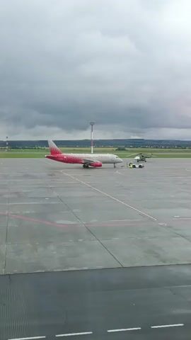 This media file shows an airport tarmac on a cloudy day. A red and white airplane with "Rossiya" branding is parked on the concrete, with a tow truck and two people in yellow vests nearby. In the background, a helicopter and a control tower are visible, suggesting an active airport environment. The overcast sky and damp tarmac indicate recent or ongoing rain. The presence of airport vehicles and personnel points to the activity of aircraft servicing and movement. The overall scene conveys a typical operational moment at an airport under subdued weather conditions.