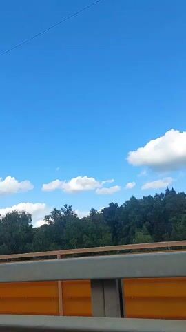 The video shows a view from a vehicle moving on a bridge. The sky is blue with scattered white clouds. On the right side of the frame, a dense forest of green trees is visible. A guardrail with a wooden top and orange panels runs along the bridge. In the distance, smoke rises from the forest on the left side of the frame. A street lamp is visible on the left side of the frame.