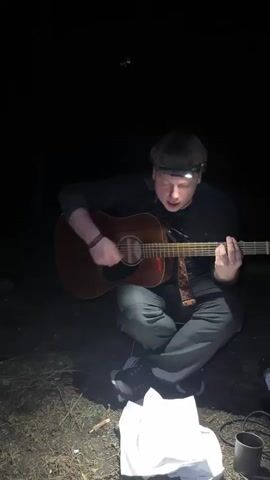 A young man sits in a darkened outdoor space, playing an acoustic guitar. He is wearing a black long-sleeved shirt, gray pants, and black sneakers. A headlamp illuminates his face. The guitar has a reddish-brown finish, and the strap is colorful. He's sitting cross-legged on the ground, which is covered in dirt and some small debris. A white paper bag and a metal mug are visible on the ground near him. The overall environment is dark, suggesting it is night.