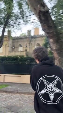 A person, seen from behind, walks along a paved sidewalk under an overcast sky. They are wearing a black hooded sweatshirt that features a large, distinct white graphic on the back: a stylized Baphomet head (a goat's head with horns) enclosed within an inverted pentagram, which is itself encircled. Smaller, similar symbols are visible on the sleeves of the sweatshirt.

The setting is a street in Olivos, Argentina. To the right of the person, a dark metal fence separates the sidewalk from a large, ornate building. This structure exhibits a Gothic Revival architectural style, characterized by its light-colored stone facade, crenellated parapets along the roofline, and arched windows. Green foliage from hedges and trees obscures parts of the building and lines the street. The sidewalk appears to be made of cobblestones or similar pavers. The lighting indicates an overcast day, with no direct sunlight creating harsh shadows. The activity shown is simply the person walking past the distinctive building. No facial expressions or discernible emotions are visible as the person's back is to the camera. There is no visible text, only the symbolic imagery on the person's clothing.