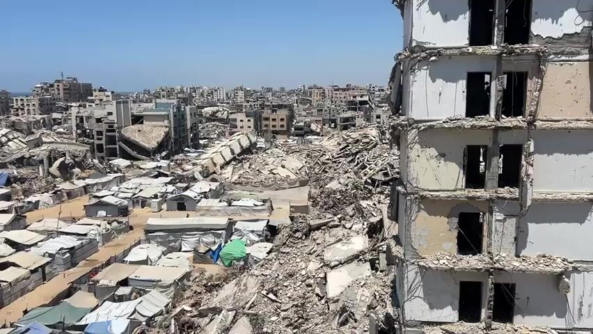 The video shows an aerial view of a city devastated by conflict. In the foreground on the right, a damaged multi-story building stands, its facade crumbling and windows gaping. To the left and center, a vast expanse of rubble fills the landscape, with collapsed buildings and debris strewn everywhere. Among the ruins, rows of makeshift tents and small structures, likely temporary shelters, are visible. In the background, more damaged buildings stretch towards the horizon, with the ocean visible in the far distance. The sky is clear and blue. There are no people clearly visible in the frame. The scene depicts widespread destruction and displacement.